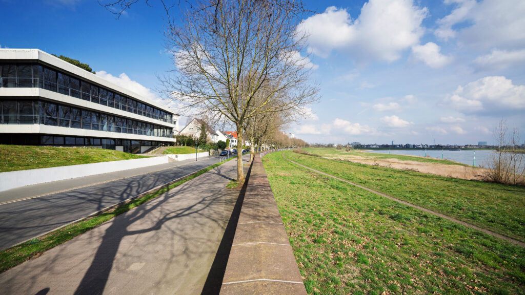 Zweistöckiges Gebäude mit Glasfassade an einer Straße, direkt daneben Deich und Fluss